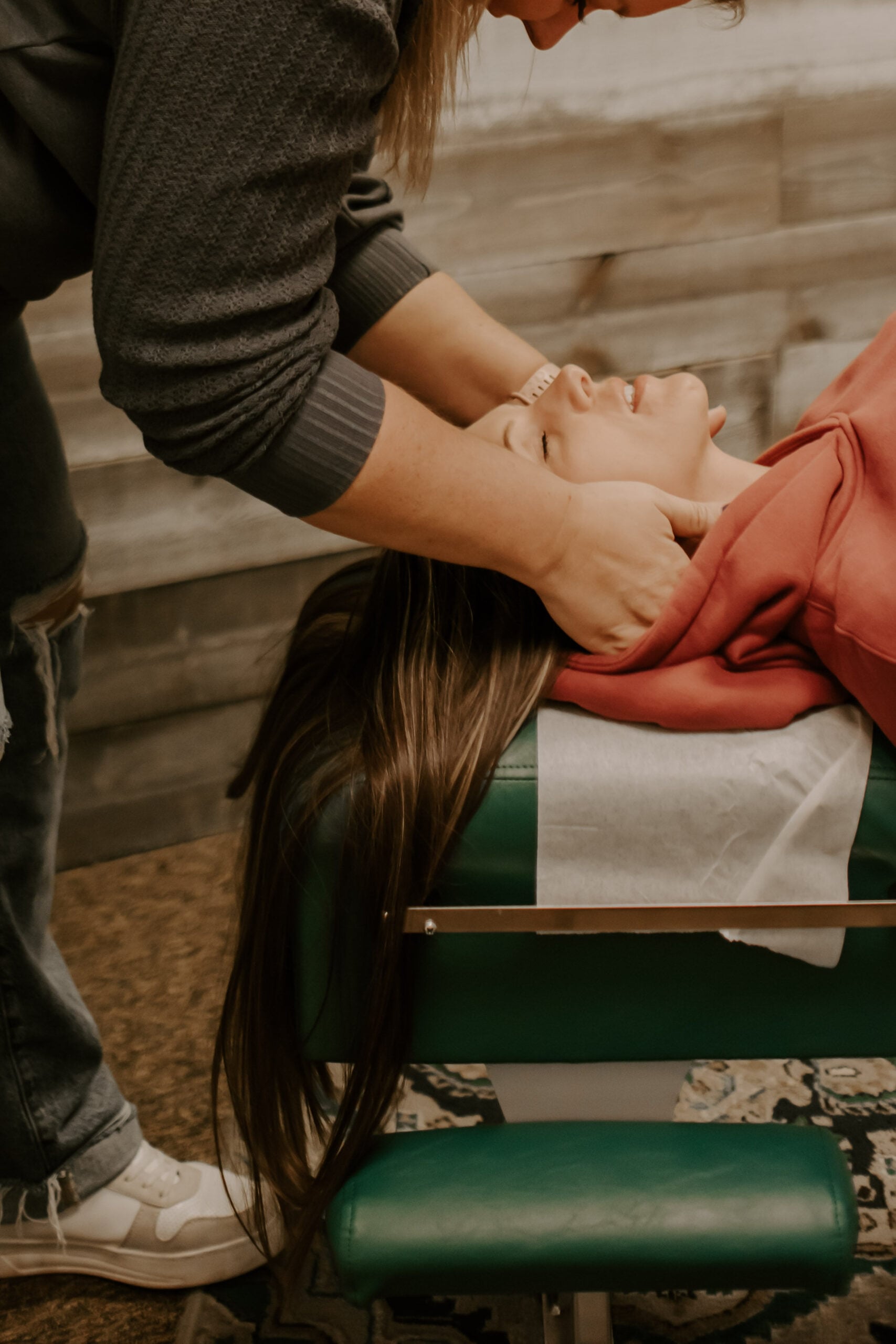 A chiropractor gently adjusts a woman’s neck while she lies on a padded green treatment table in a clinic setting.