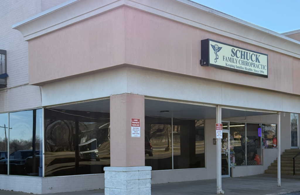 Exterior of Schuck Family Chiropractic office in a strip mall, featuring a beige and light pink corner storefront with large tinted windows, a covered walkway, and a sign reading “Schuck Family Chiropractic – Keeping Families Healthy Since 1994” mounted near the roofline.